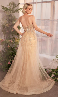 A woman with short blonde hair wears the Andrea & Leo A1365 Strapless Sequin Prom Gown, a floor-length dress with a sheer, embroidered back, standing before a window framed by greenery and flowers.