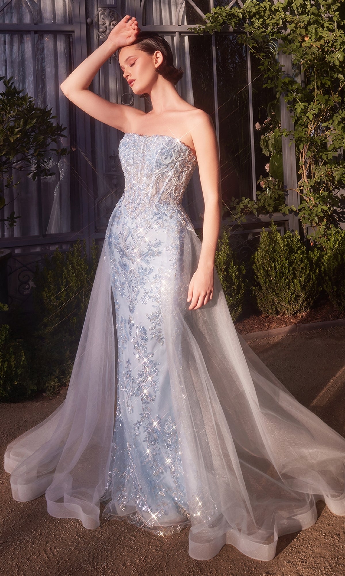 A woman stands outdoors in the sunlight, wearing the Andrea & Leo A1365 strapless sequin prom gown, featuring light blue embroidery and a sheer train.