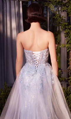 A person stands outside, back facing, wearing the Strapless Sequin Prom Gown: Andrea & Leo A1365 in light blue, featuring a lace-up back, sheer tulle, and floral details. Shrubs and a window are visible in the background.