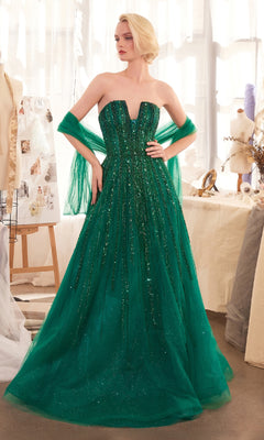 A woman in the Andrea & Leo A1369 Strapless Beaded Long Prom Dress, floor-length and emerald green with glitter details, stands in a well-lit studio draped with fabric and dressmaking materials.