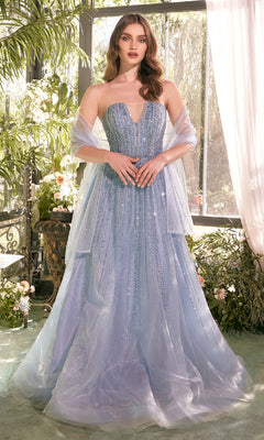 A woman wearing the Andrea & Leo A1369 strapless beaded long prom dress with a sheer shawl stands in a sunlit conservatory, surrounded by greenery and flowers.