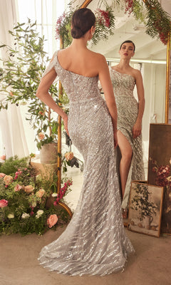 A woman in the Andrea & Leo A1370 long formal dress, featuring a silver beaded design and asymmetrical neckline, stands before a mirror amid flowers and greenery in a softly lit room.