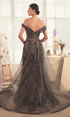 A woman stands in a studio, facing away, wearing the Long Dark Silver Formal Dress: Andrea & Leo A1375 with beaded details and a tulle train. Sketches and a dress form in the background highlight its elegant, classic prom style.