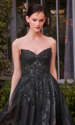 A woman in the Andrea & Leo A1378 strapless long prom dress with a sweetheart neckline, corset waist, and embroidered floral lace stands by a glass window surrounded by greenery.