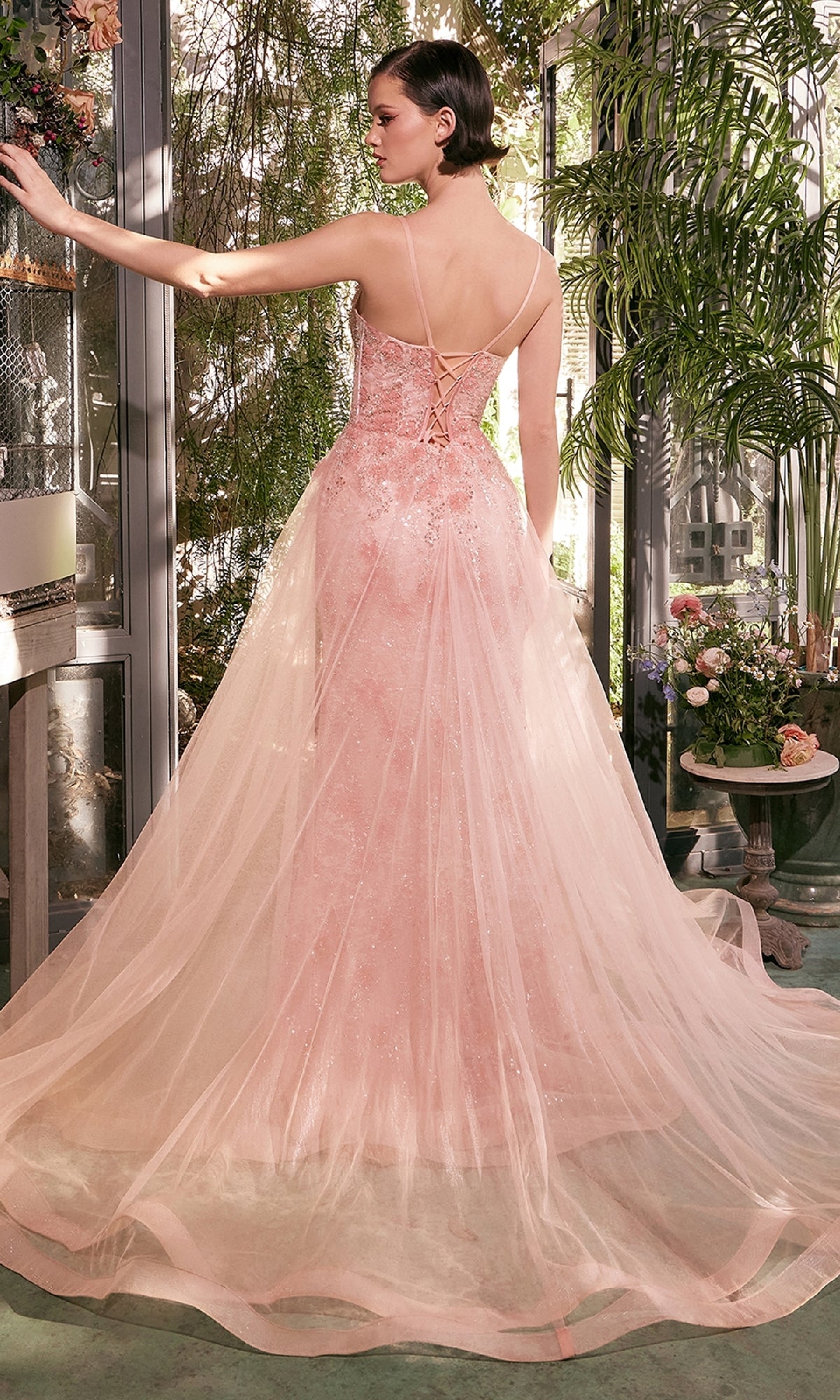 A woman wearing the Andrea and Leo A1382 Long Glitter Prom Dress—a light pink, strapless, floor-length gown with a corset bodice and tulle overlay—stands indoors near greenery and flowers.