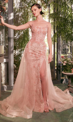 A woman stands indoors in the Andrea and Leo A1382 long glitter prom dress, featuring blush pink fabric, floral embroidery, a thigh-high slit, and a tulle overlay, surrounded by greenery and flowers.
