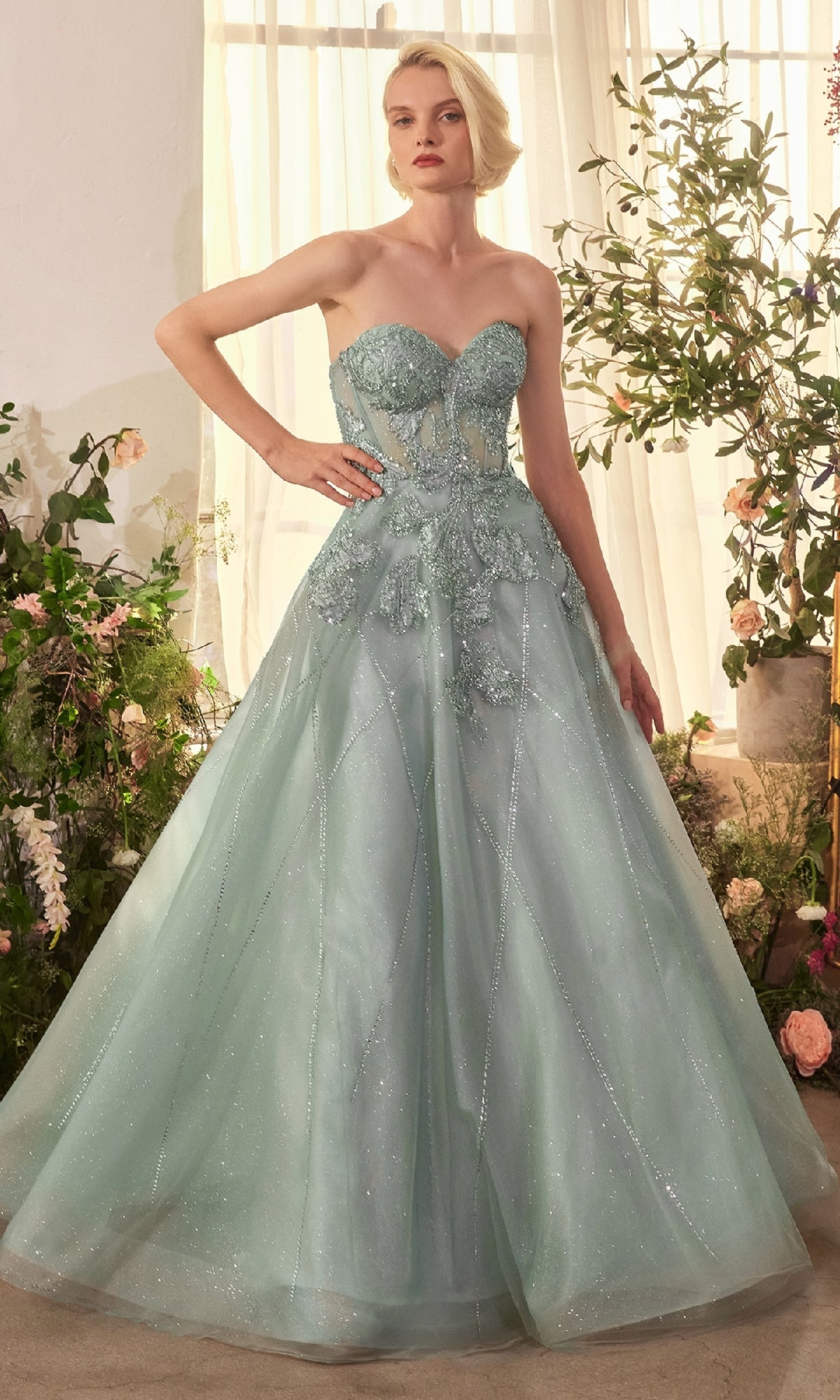 A woman stands indoors among flowers, wearing the Andrea & Leo A1387 Strapless Long Prom Ball Gown in light blue with a beaded bodice and full shimmery skirt—a stunning A-line dress perfect for any special occasion.