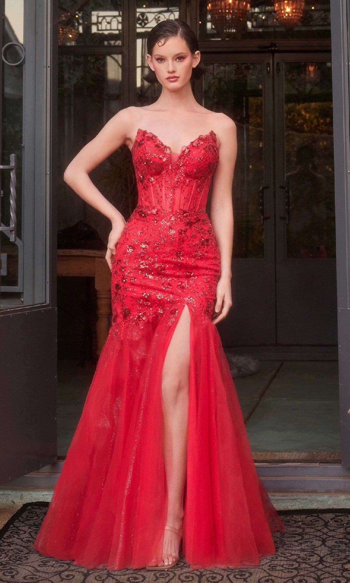 Woman wearing the Andrea & Leo A1388 strapless mermaid prom dress with floral embellishments and a thigh-high slit, standing before large glass doors.