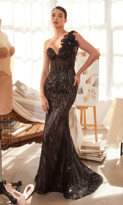 A woman stands indoors wearing the Andrea & Leo A1392, a one-shoulder beaded prom gown—strapless, floor-length, black with sequin details and a floral accent on one shoulder.
