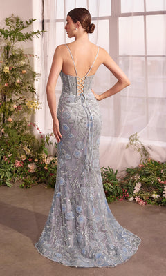 A woman stands indoors near large windows and greenery, wearing the Blue Floral Long Prom Dress: Andrea & Leo A1393—a light blue, floor-length mermaid gown featuring floral embroidery and a lace-up back that captures timeless elegance.