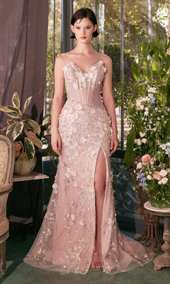 A woman stands indoors wearing the Strapless Floral Prom Gown: Andrea & Leo A1396, a blush pink lace dress with a thigh-high slit; plants and flowers decorate the background.