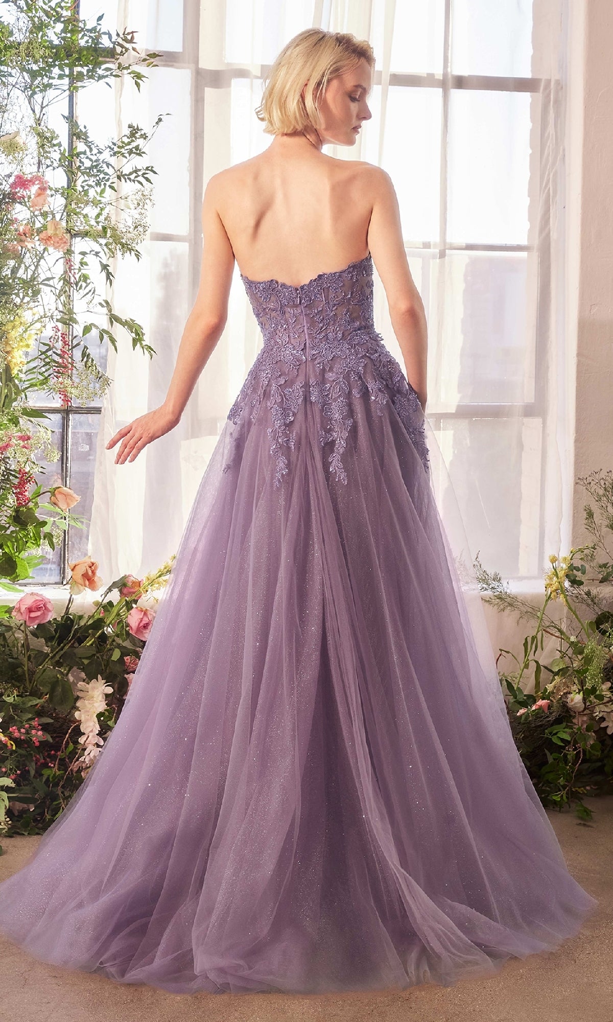 A woman stands indoors by a window wearing the Andrea & Leo A1398 Strapless Glitter Prom Gown with lace detailing and a tulle skirt; flowers are arranged nearby.