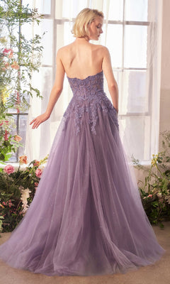 A woman stands indoors by a window wearing the Andrea & Leo A1398 Strapless Glitter Prom Gown with lace detailing and a tulle skirt; flowers are arranged nearby.
