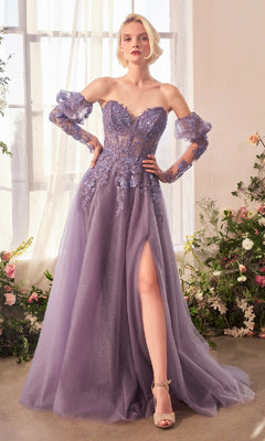 A woman stands in a room wearing the Andrea & Leo A1398 Strapless Glitter Prom Gown, a long violet dress with sparkle accents and a thigh-high slit. Flowers and natural light fill the background.