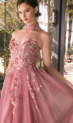 A person wearing the Andrea & Leo A1399 Strapless Floral Prom Gown in pink, featuring embroidered floral applique and a matching choker, stands near a glass door in a sunlit room.