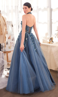 A woman stands in a sunlit room wearing the Strapless Floral Prom Gown: Andrea & Leo A1399, a blue, floor-length dress with floral appliqué and lace-up back details.
