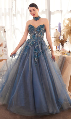 A woman models the Andrea & Leo A1399 Strapless Floral Prom Gown—an elegant blue ball gown with floral applique and a tulle skirt—in a bright studio filled with dress forms and artistic materials.