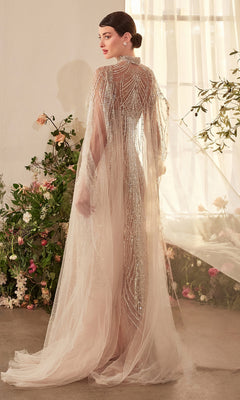 A woman stands indoors near flowers, wearing the Andrea and Leo A1411 long formal dress with a beaded design, lace-up back, and removable cape. Facing away, she highlights the gown’s intricate details as light filters through a window.
