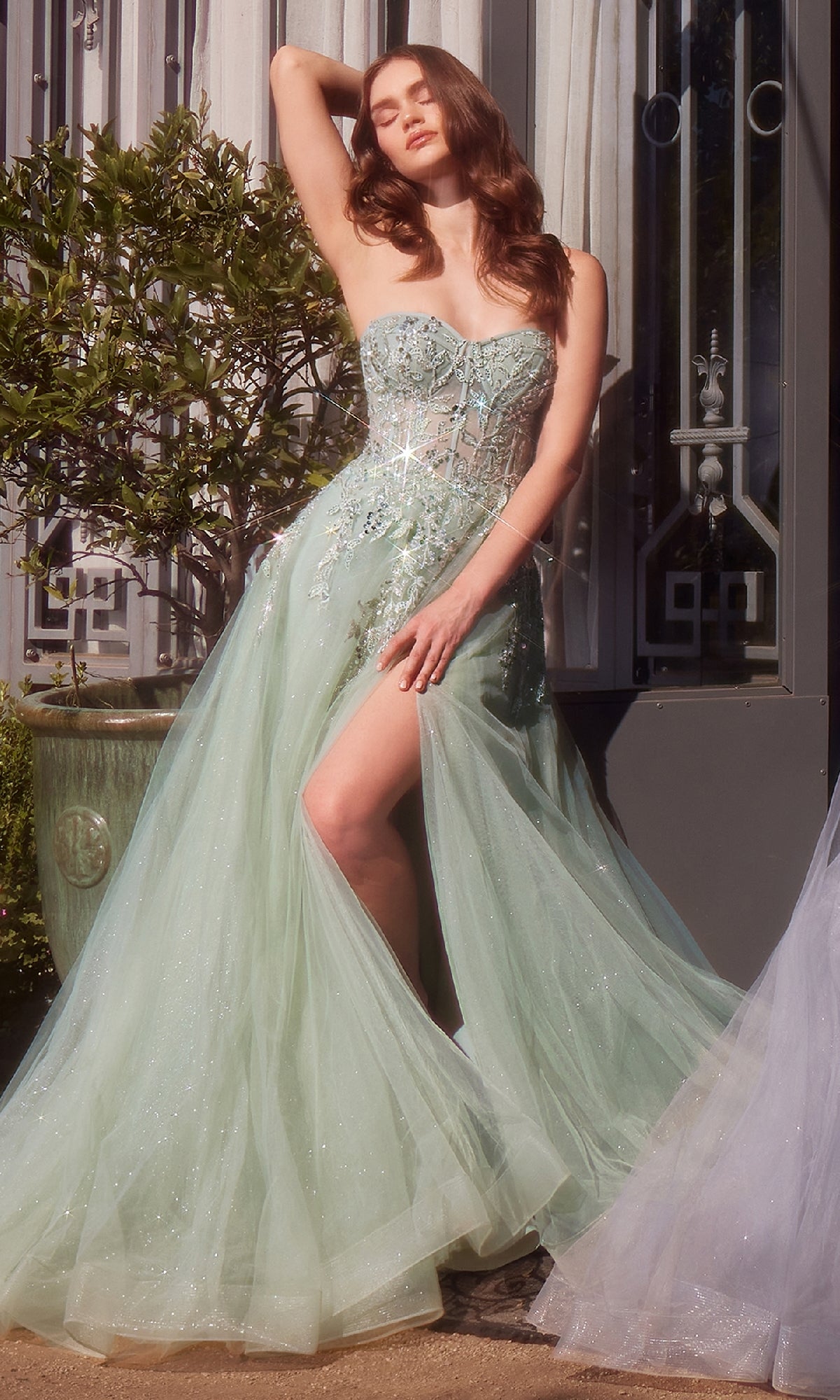 A woman in a sheer, light green Strapless Prom Ball Gown: Andrea & Leo A1415 with floral embellishments stands outdoors, posing with one arm raised and revealing one leg through a thigh-high slit.