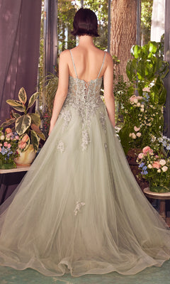 A woman with a short bob wears the Andrea & Leo A1417 Embroidered Long Prom Dress—an elegant light green gown with an embroidered bodice and tulle train—standing indoors among flowers, facing away from the camera.