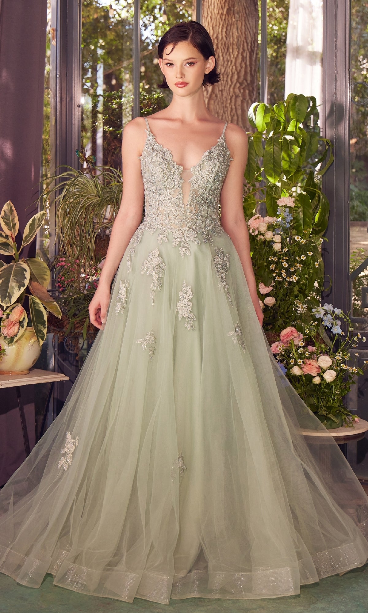 A woman stands indoors in the Andrea & Leo A1417 embroidered long prom dress, surrounded by various potted plants and flowers.