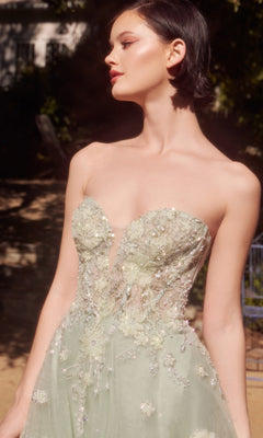 A woman stands outdoors in sunlight wearing the Sage Green Prom Ball Gown: Andrea & Leo A1418, a strapless corset dress adorned with floral embroidery and beading.