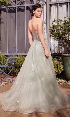 A woman wears the Andrea & Leo A1418 sage green prom ball gown with floral embellishments. The strapless corset dress has a lace-up back and sweeping train as she stands outdoors, glancing over her shoulder.