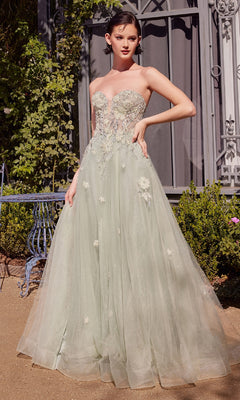 A woman stands outdoors on a gravel path near lush greenery and an ornate wrought iron window, wearing the Sage Green Prom Ball Gown: Andrea & Leo A1418, a strapless corset gown with floral embroidery.