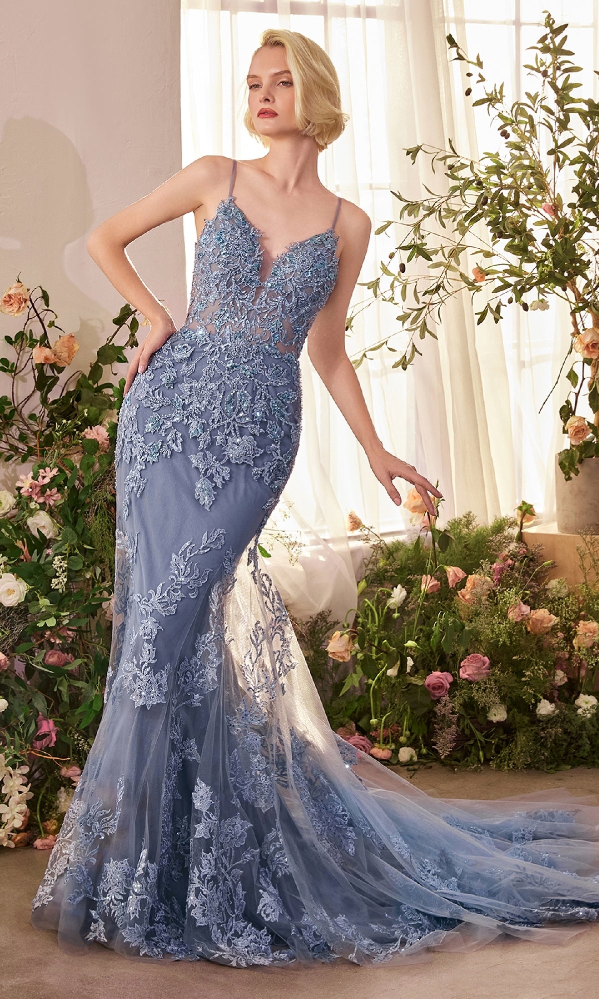 A woman in the Beaded-Lace Long Prom Dress Andrea & Leo A1421 stands by a window, surrounded by flowers and greenery. The fitted, floor-length gown features blue beaded lace with elegant floral embroidery.