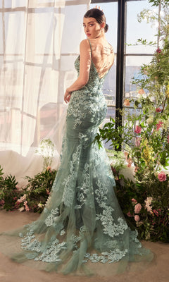 A woman stands indoors near large windows and flowers, wearing the Andrea & Leo A1421 Beaded-Lace Long Prom Dress, a form-fitting light blue-green gown with sheer back, beaded lace details, and a dramatic train.