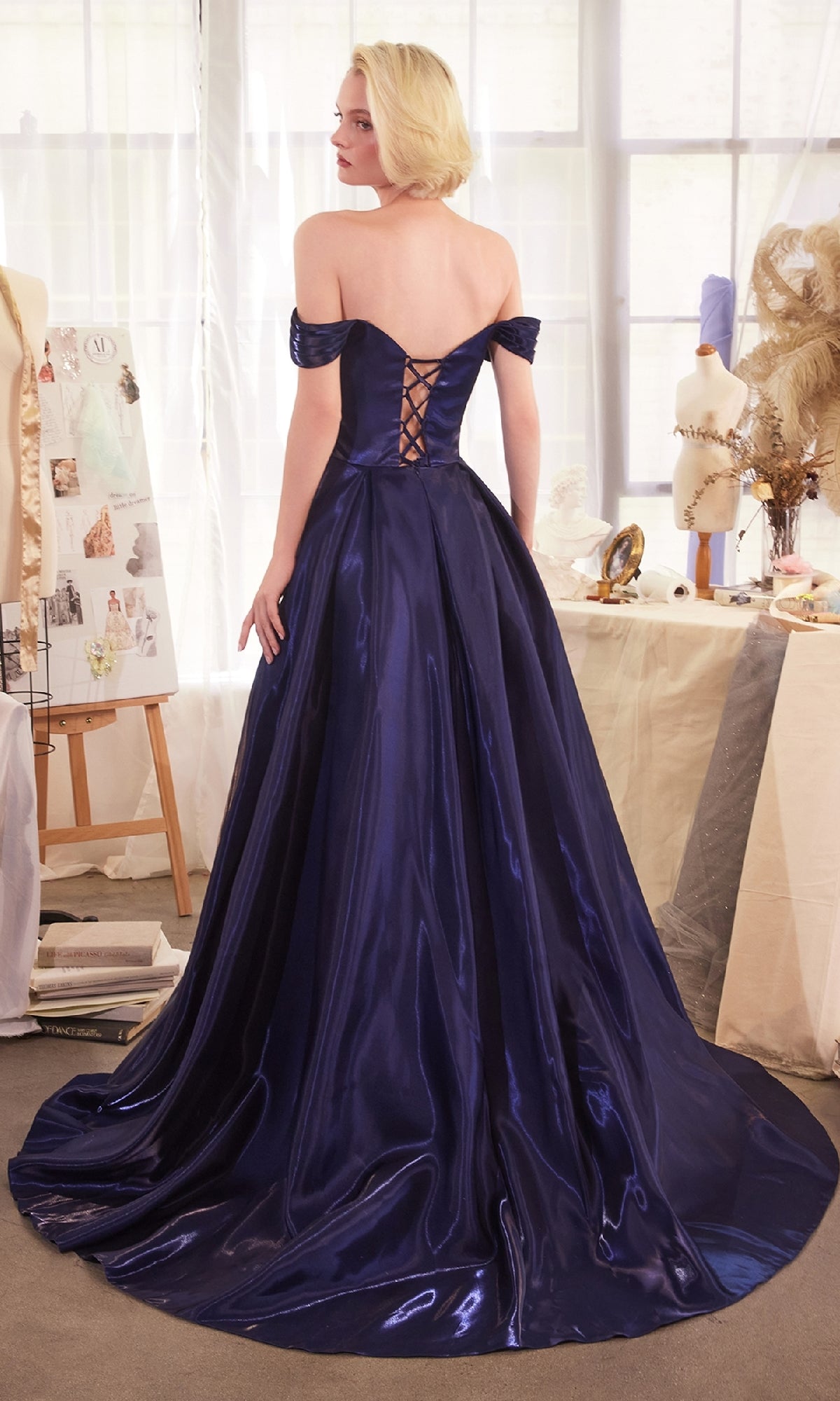 A person wearing the Off-the-Shoulder Prom Ball Gown: Andrea & Leo A1424, a floor-length dark blue dress with a lace-up back, stands in a room filled with mannequins, dried flowers, and fashion sketches.