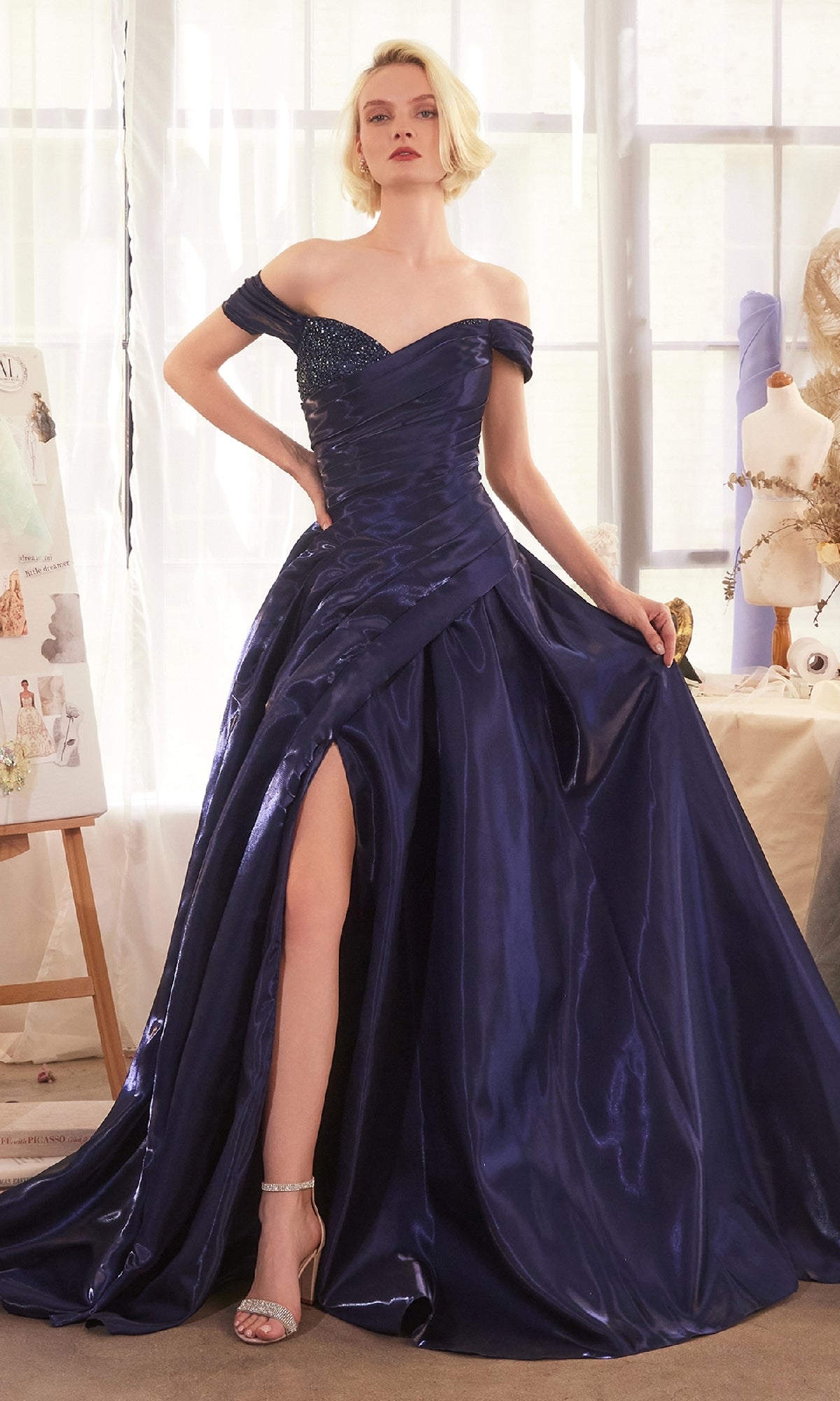 A woman in the Off-the-Shoulder Prom Ball Gown: Andrea & Leo A1424, a dark blue A-line evening dress with a thigh-high slit, stands indoors near a window and mannequins, holding the skirt of her elegant gown.
