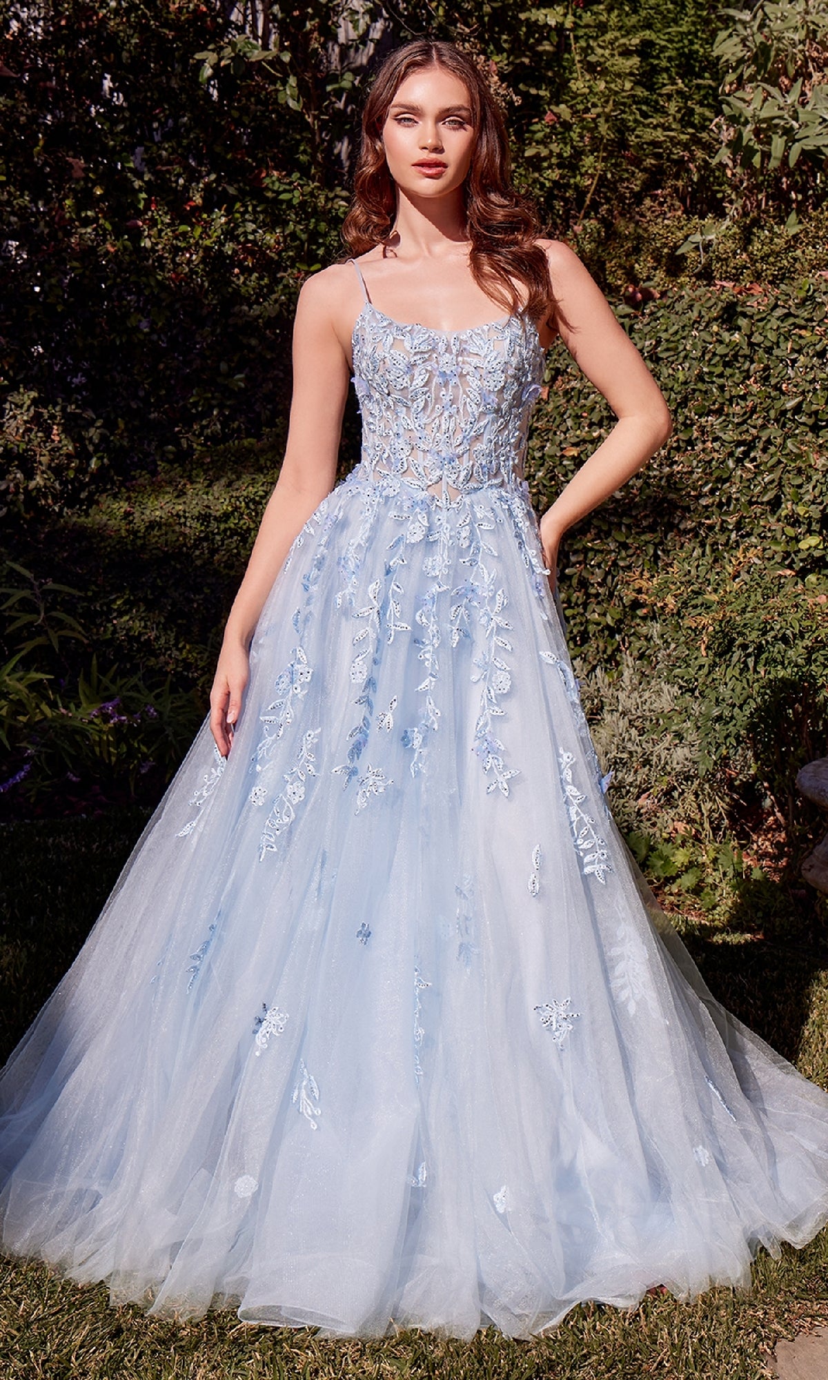 A woman stands outdoors in the Andrea and Leo A1437 long blue prom ball gown, featuring floral embroidery and a tulle skirt that beautifully complements the surrounding green foliage and grass.