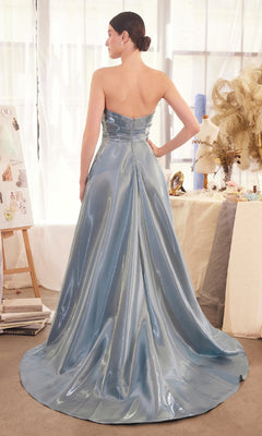 A woman stands indoors wearing the Andrea & Leo A1439 long prom dress—a strapless blue gown with an a-line skirt and flowing train, its liquid satin fabric catching the light. She faces away, surrounded by mannequins and art supplies.