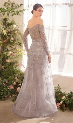 A woman stands indoors by a window, surrounded by greenery and flowers, wearing the shimmering Andrea and Leo A1445 long formal dress. This off-the-shoulder tulle gown adds elegance to the enchanting scene.