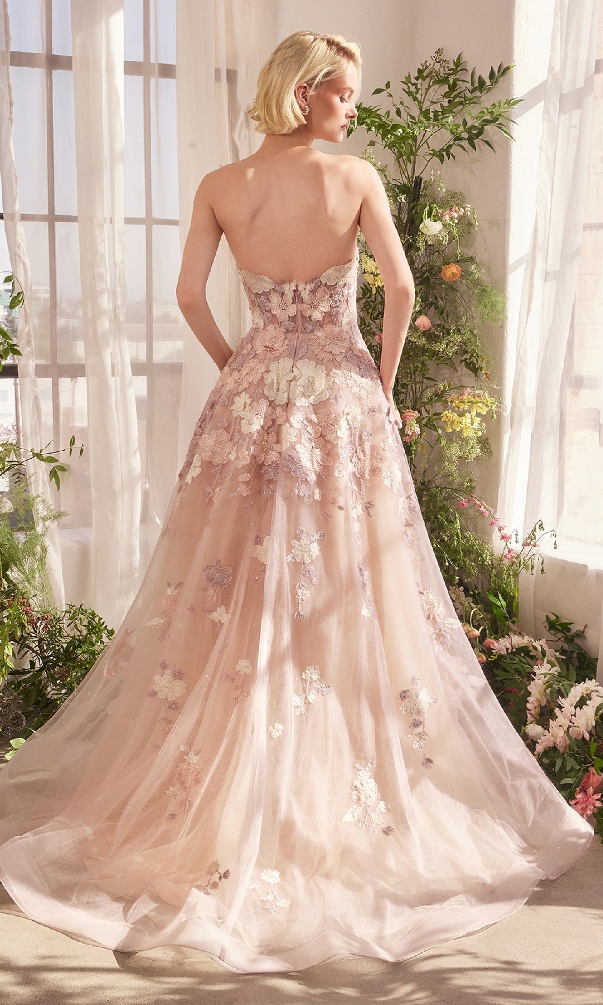 A woman stands indoors near large windows, surrounded by plants and flowers, wearing the Strapless Long Mauve Prom Gown: Andrea & Leo A1453 with a flowing train.