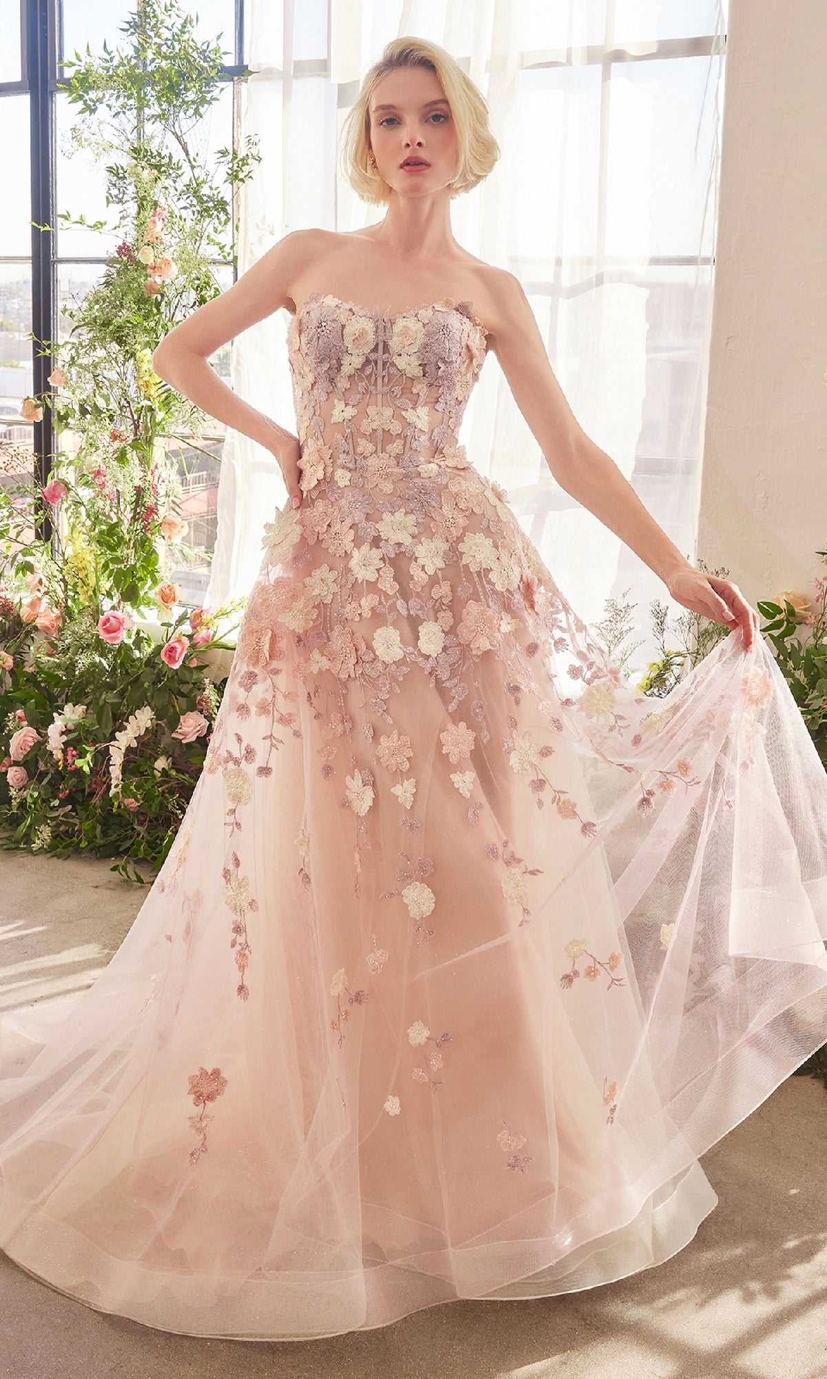 A woman stands indoors in the Andrea & Leo A1453 Strapless Long Mauve Prom Gown with floral appliqués and a sheer tulle skirt, surrounded by flowers and sunlight.
