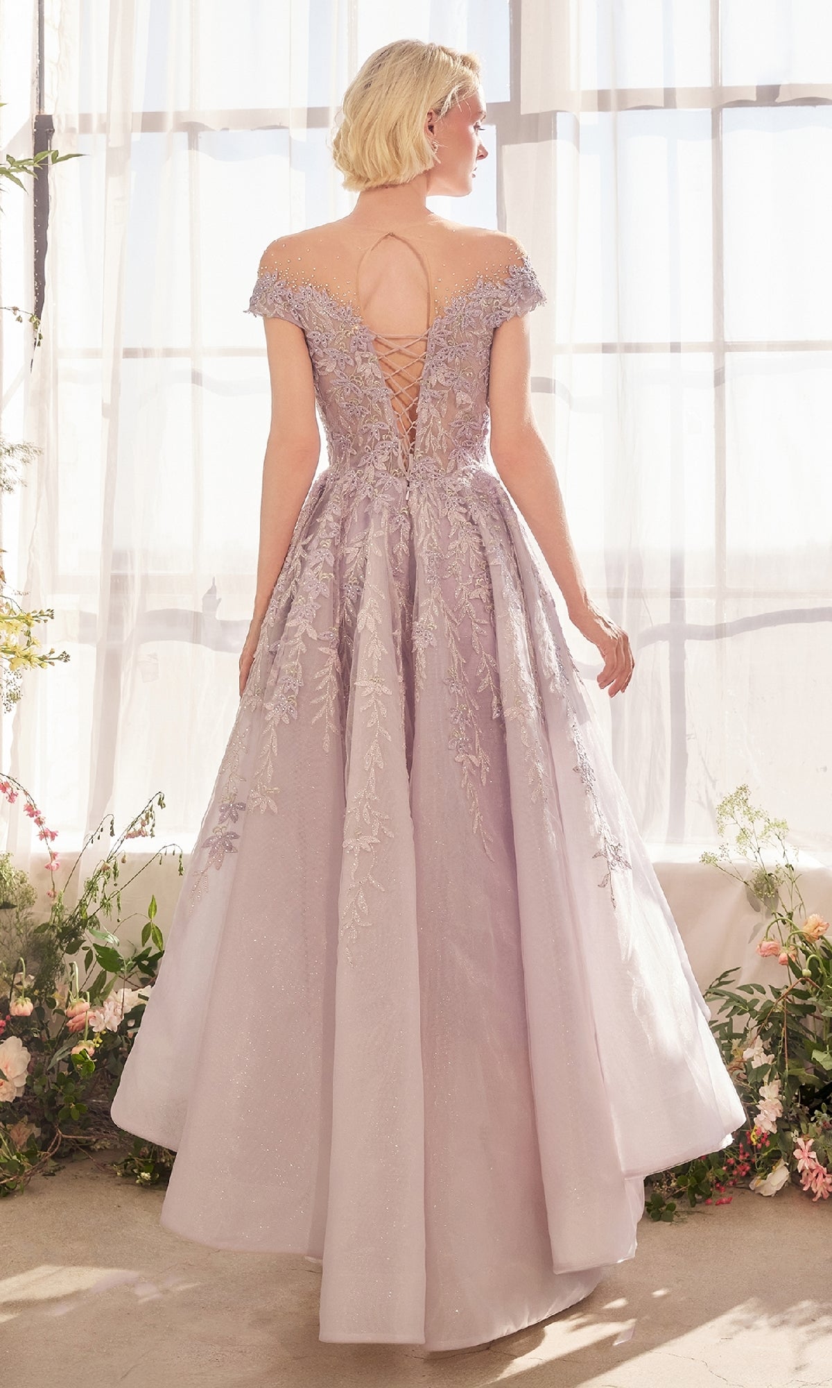 A woman wears the Andrea and Leo A1454 long prom dress featuring pastel tones, floral embroidery, and a lace-up back, standing before sheer curtains and floral decorations.