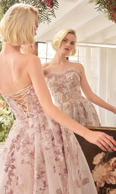 A woman with softly waved blonde hair stands before a mirror, her hand on the frame, wearing the Andrea and Leo A1455 long prom dress—a strapless organza gown featuring floral lace embroidery.