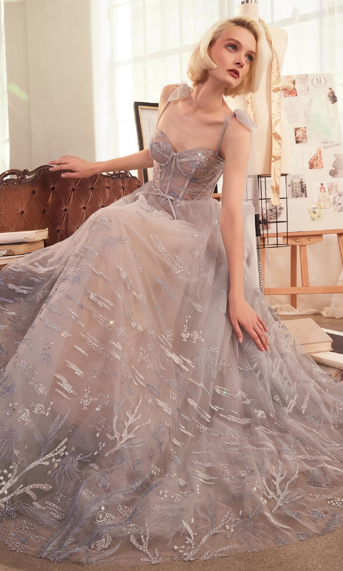 A woman in the Andrea & Leo A1458 long silver prom ball gown sits on a sofa in a well-lit room, surrounded by dress sketches in the background.