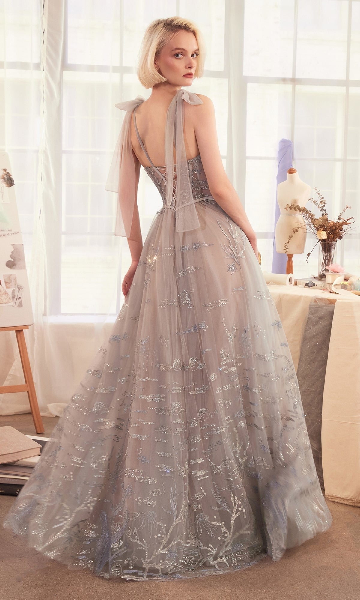 A woman in the Andrea & Leo A1458 long silver prom ball gown stands in a softly lit room, looking over her shoulder.