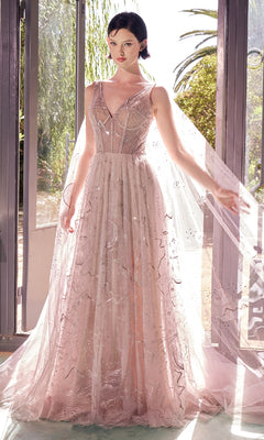 A woman stands indoors in the Andrea & Leo A1459 Caped Long Nude Prom Dress, featuring sheer fabric, beaded details, and a flowing cape, illuminated by sunlight through large glass doors behind her.