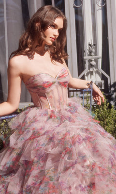 A woman in the Andrea & Leo A1463 Strapless Print Prom Ball Gown with a sweetheart corset bodice and layered tulle skirt sits on a metal chair outdoors near a window.