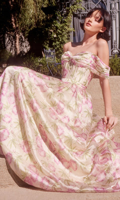 A woman sits outdoors in the Andrea & Leo A1466 long floral-print prom dress, its off-the-shoulder design, light pink and green blooms, and corset-style bodice beautifully highlighted by the sunlight.