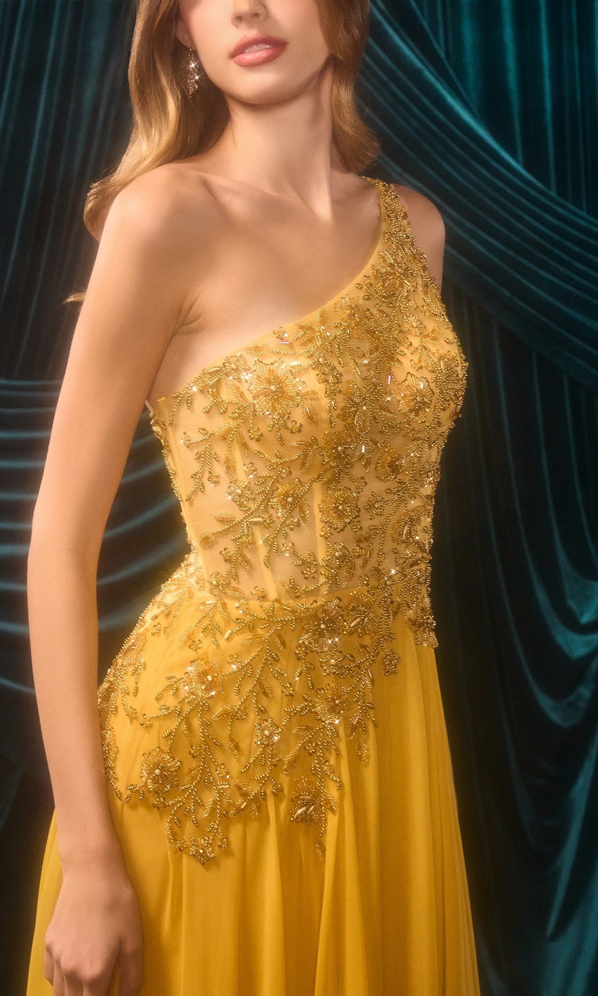 A woman in the Andrea & Leo A1495 One-Shoulder Yellow Prom Gown, adorned with intricate gold embroidery, stands before dark green velvet draped curtains.