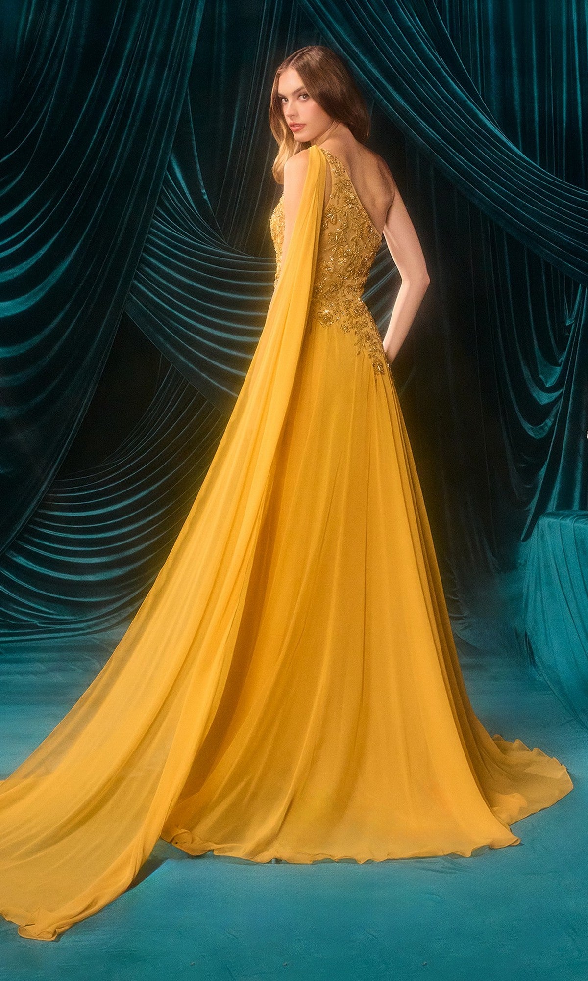 A woman in the Andrea & Leo A1495 One-Shoulder Yellow Prom Gown stands against a backdrop of dark teal draped fabric.
