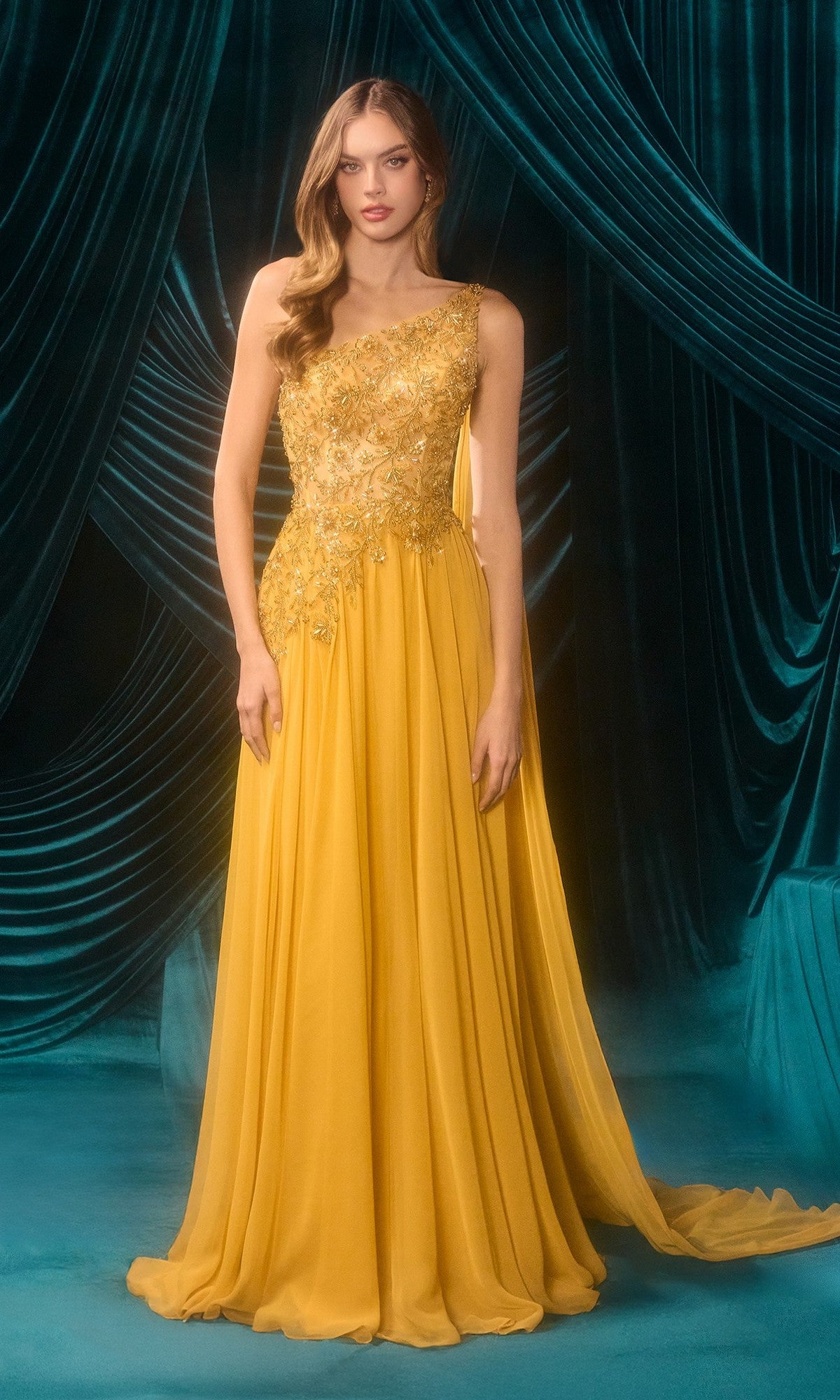A woman stands before dark teal drapes in the Andrea & Leo A1495 One-Shoulder Yellow Prom Gown in marigold, featuring floral embroidery and a flowing, floor-length silhouette.