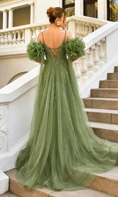 A woman stands on outdoor stairs, facing away, wearing the Andrea & Leo A1515 long prom dress—an elegant, floor-length green gown featuring floral sleeve accents and a lace-up back. The look is enhanced by a detachable tulle cape that adds an ethereal touch of grace.