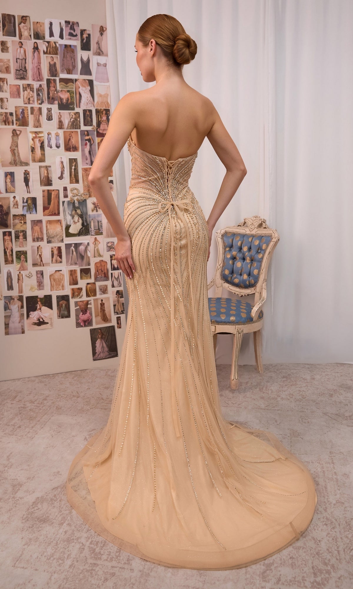A woman wearing the Andrea & Leo A1519 long prom dress, featuring a fitted silhouette, strapless sweetheart neckline, and a beige hue with a dramatic train, stands facing a wall covered in fashion photos beside a blue and gold chair.