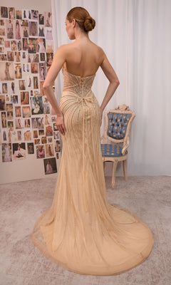 A woman wearing the Andrea & Leo A1519 long prom dress, featuring a fitted silhouette, strapless sweetheart neckline, and a beige hue with a dramatic train, stands facing a wall covered in fashion photos beside a blue and gold chair.